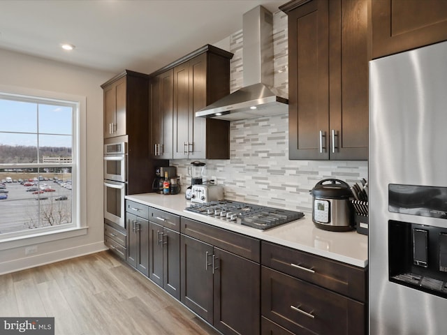 kitchen featuring decorative backsplash, dark brown cabinetry, stainless steel appliances, wall chimney range hood, and light hardwood / wood-style flooring