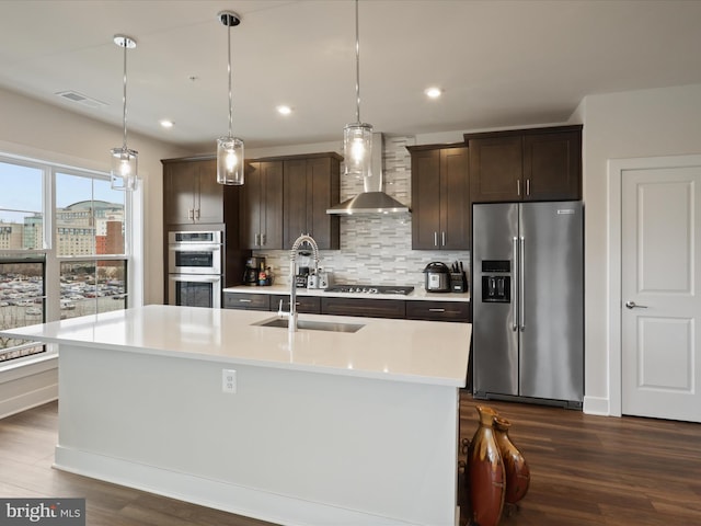 kitchen with an island with sink, sink, hanging light fixtures, stainless steel appliances, and wall chimney range hood