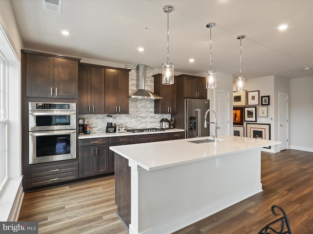 kitchen featuring decorative light fixtures, an island with sink, sink, stainless steel appliances, and wall chimney range hood