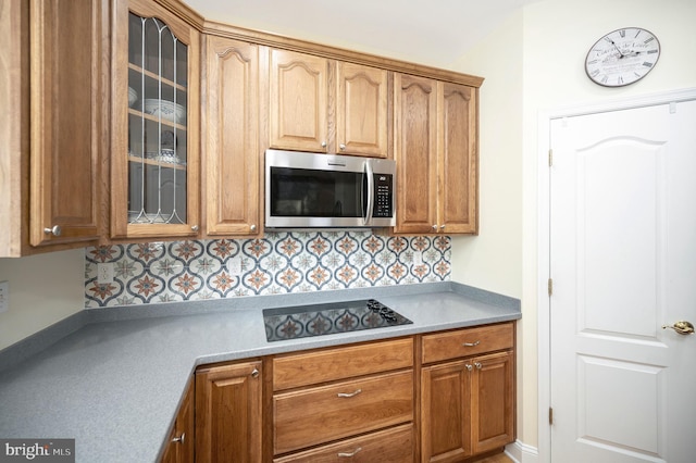 kitchen with glass insert cabinets, stainless steel microwave, brown cabinets, black electric stovetop, and backsplash