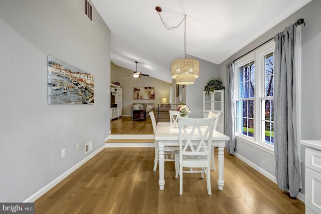 dining area featuring ceiling fan with notable chandelier, wood finished floors, visible vents, baseboards, and vaulted ceiling