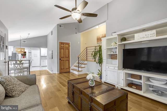 living area featuring light wood finished floors, visible vents, a ceiling fan, stairway, and high vaulted ceiling