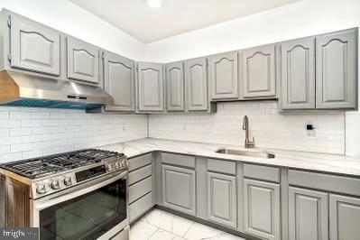 kitchen with stainless steel range with gas cooktop, gray cabinets, a sink, under cabinet range hood, and tasteful backsplash