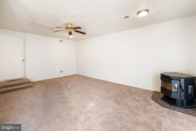 carpeted spare room featuring a textured ceiling, a ceiling fan, visible vents, attic access, and a wood stove