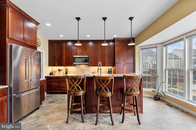 kitchen with a center island with sink, decorative backsplash, appliances with stainless steel finishes, light stone counters, and hanging light fixtures