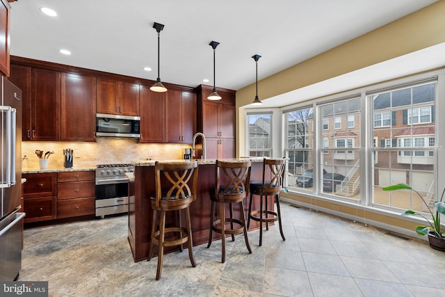 kitchen with decorative light fixtures, tasteful backsplash, appliances with stainless steel finishes, a kitchen island with sink, and light stone countertops