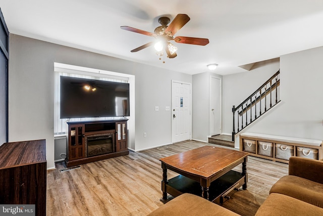 living room featuring baseboards, stairs, wood finished floors, a glass covered fireplace, and a ceiling fan