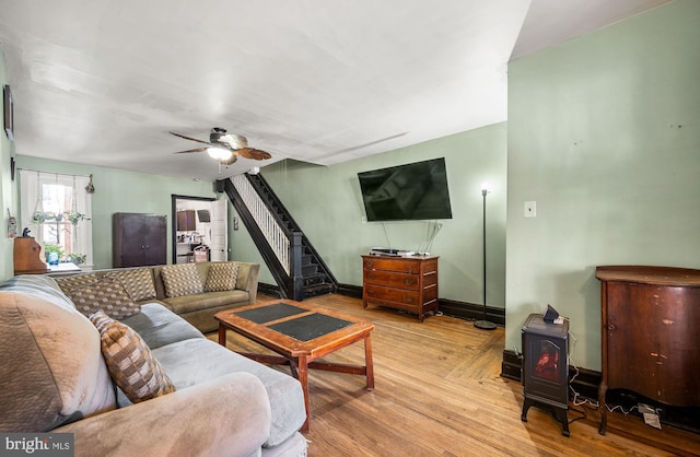 living room featuring baseboards, a ceiling fan, light wood-style flooring, a wood stove, and stairs