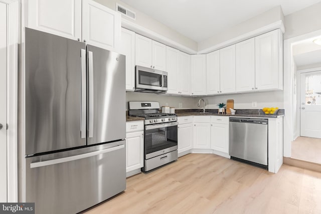 kitchen with appliances with stainless steel finishes, sink, dark stone countertops, white cabinets, and light hardwood / wood-style floors