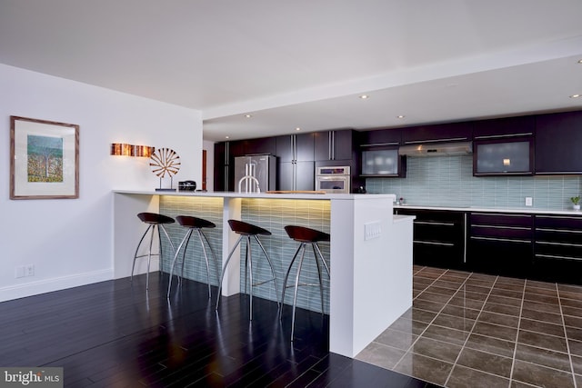 kitchen featuring a breakfast bar area, appliances with stainless steel finishes, dark wood-type flooring, decorative backsplash, and dark brown cabinetry