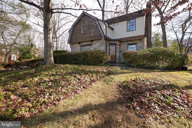 view of front facade with stone siding, a chimney, a front yard, and a gambrel roof