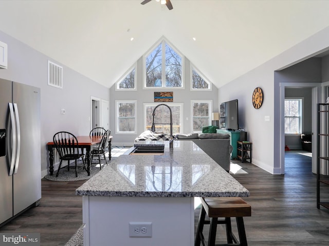 kitchen with dark wood-type flooring, light stone counters, high vaulted ceiling, stainless steel fridge, and a kitchen island with sink