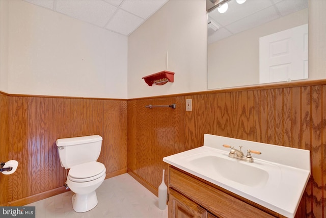 half bathroom with wainscoting, toilet, vanity, a paneled ceiling, and wood walls