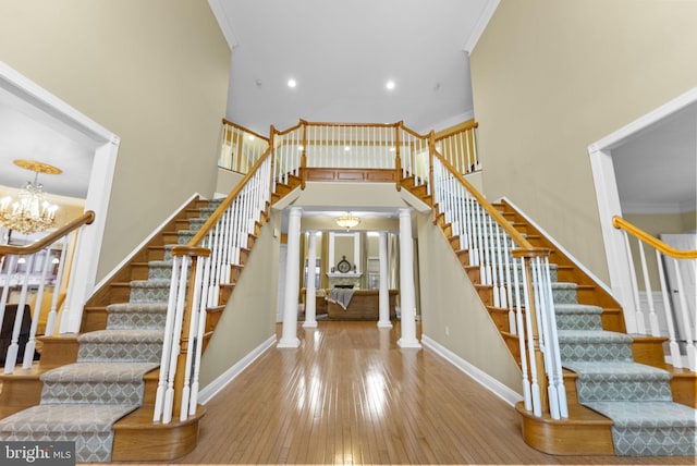 staircase with crown molding, hardwood / wood-style flooring, an inviting chandelier, a towering ceiling, and decorative columns