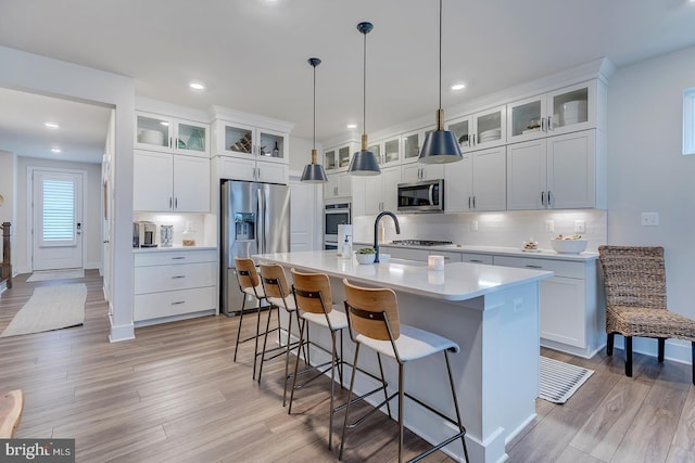 kitchen featuring white cabinets, glass insert cabinets, a kitchen island with sink, stainless steel appliances, and light countertops