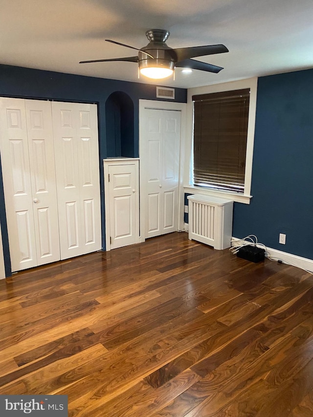 unfurnished bedroom featuring dark hardwood / wood-style flooring, two closets, radiator heating unit, and ceiling fan