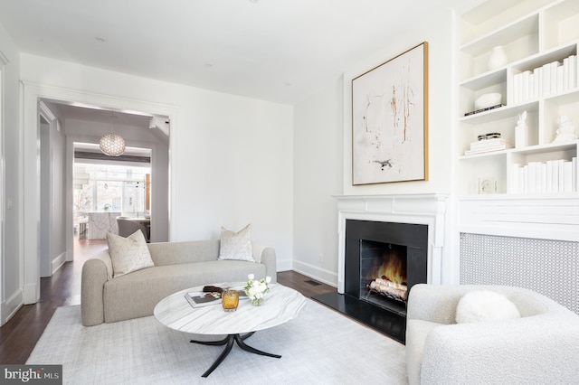 living room featuring built in features, baseboards, a warm lit fireplace, and dark wood finished floors