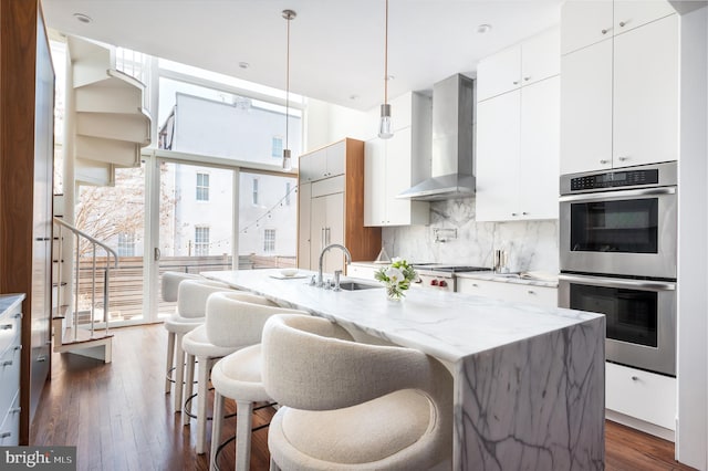 kitchen with dark wood-style floors, a sink, decorative backsplash, double oven, and wall chimney range hood