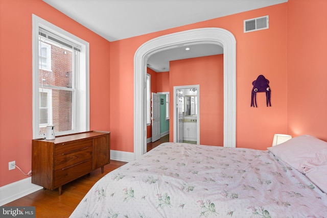 bedroom with arched walkways, visible vents, ensuite bathroom, wood finished floors, and baseboards