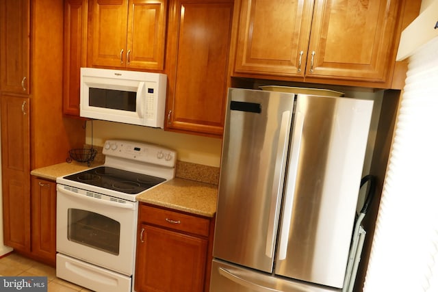 kitchen featuring brown cabinetry, light tile patterned floors, white appliances, and light stone countertops