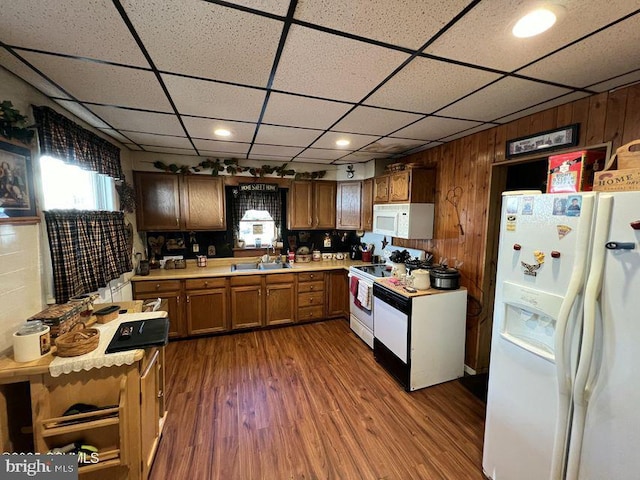 kitchen with sink, white appliances, dark wood-type flooring, wooden walls, and a drop ceiling