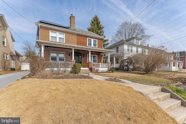 american foursquare style home featuring a front lawn, covered porch, brick siding, solar panels, and a chimney