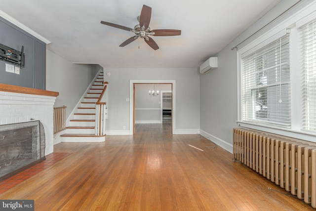 unfurnished living room featuring a wall unit AC, stairway, radiator heating unit, a fireplace, and ceiling fan with notable chandelier