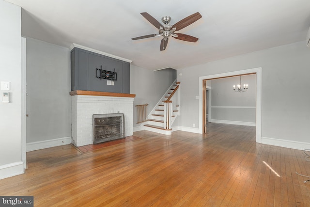 unfurnished living room featuring ceiling fan with notable chandelier, light wood finished floors, baseboards, a brick fireplace, and stairs