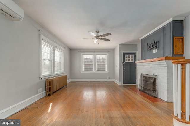 unfurnished living room featuring a wall mounted air conditioner, a ceiling fan, radiator, light wood finished floors, and a brick fireplace