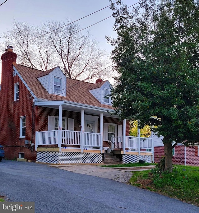 cape cod house with covered porch, roof with shingles, brick siding, and a chimney