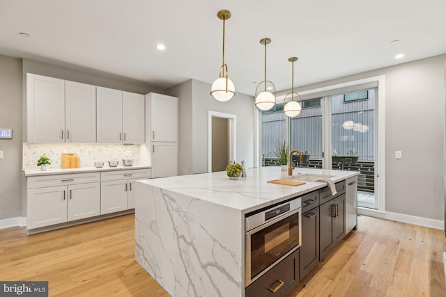 kitchen featuring tasteful backsplash, white cabinets, hanging light fixtures, a kitchen island with sink, and light stone counters