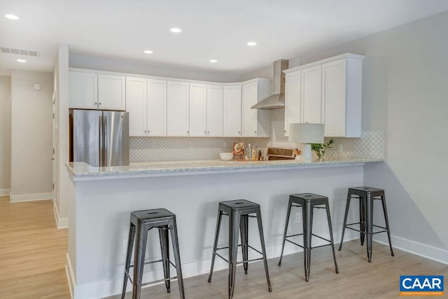 kitchen featuring wall chimney range hood, light hardwood / wood-style flooring, stainless steel fridge, a breakfast bar, and white cabinets