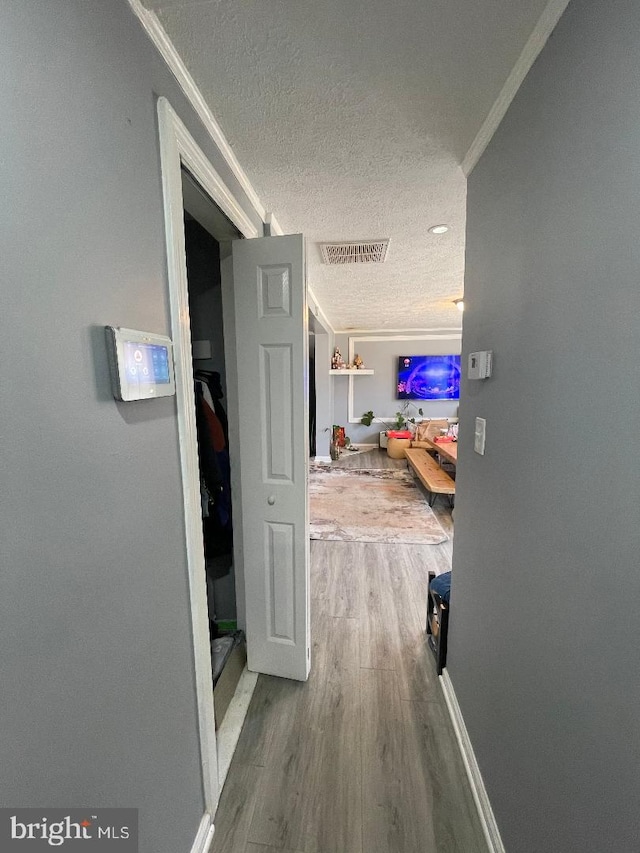 hallway with a textured ceiling, wood finished floors, visible vents, baseboards, and ornamental molding