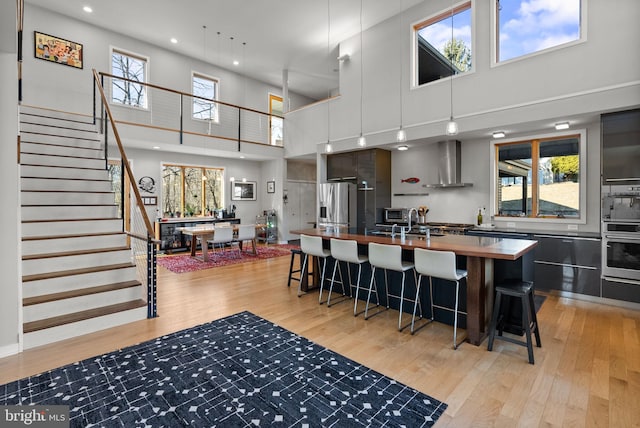 kitchen with stainless steel fridge with ice dispenser, a center island with sink, butcher block counters, light wood-type flooring, and wall chimney exhaust hood