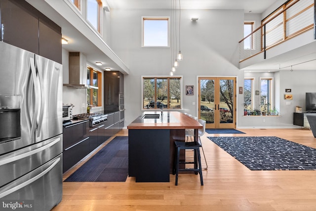 kitchen with a sink, wood counters, dark cabinetry, stainless steel appliances, and wall chimney range hood