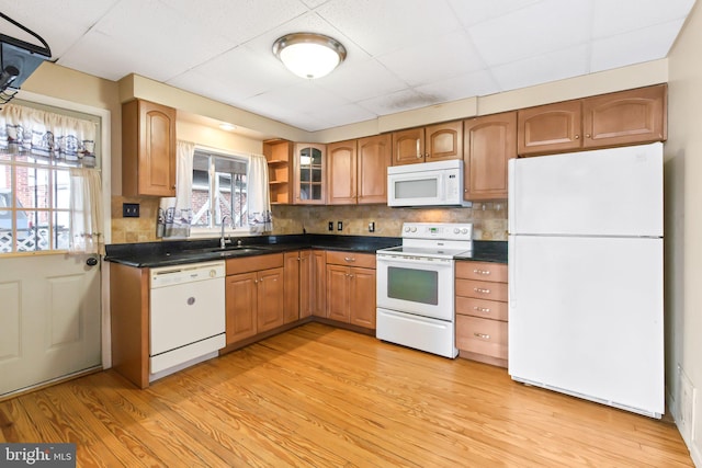 kitchen with white appliances, a sink, brown cabinets, dark countertops, and glass insert cabinets