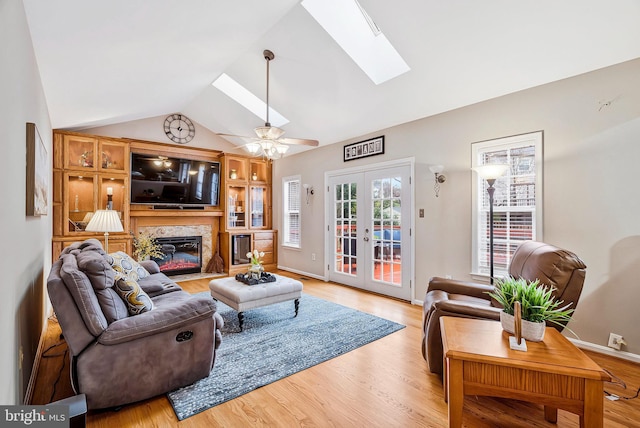 living area featuring french doors, a fireplace, light wood-style flooring, vaulted ceiling with skylight, and baseboards