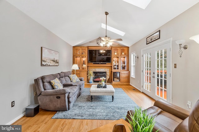 living room with vaulted ceiling with skylight, a ceiling fan, a glass covered fireplace, light wood-style flooring, and french doors