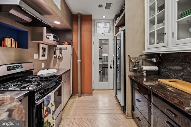 kitchen featuring stainless steel gas range oven, dark countertops, glass insert cabinets, and range hood