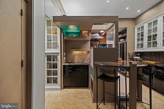 kitchen featuring glass insert cabinets, freestanding refrigerator, white cabinetry, wall chimney range hood, and dark stone counters