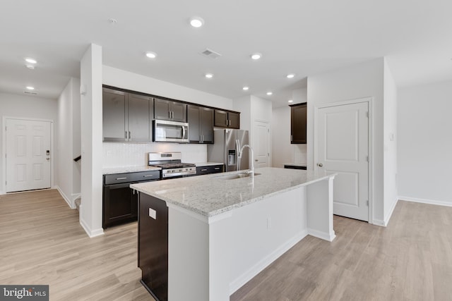 kitchen with appliances with stainless steel finishes, a kitchen island with sink, light stone counters, and light hardwood / wood-style floors