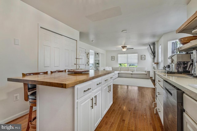 kitchen featuring stainless steel dishwasher, wood finished floors, butcher block countertops, a breakfast bar, and white cabinetry