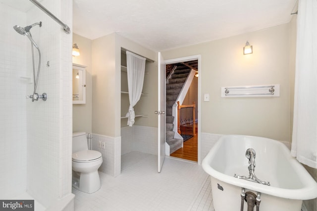 full bath featuring tile patterned flooring, toilet, a freestanding tub, tile walls, and wainscoting