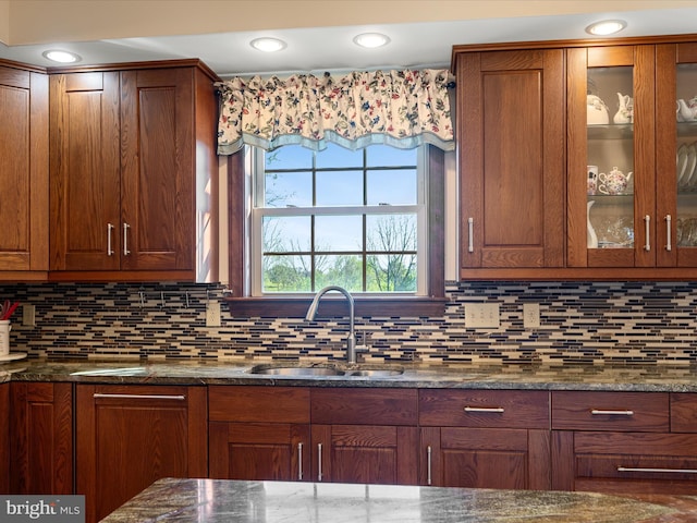 kitchen featuring a sink, glass insert cabinets, backsplash, and dark stone counters