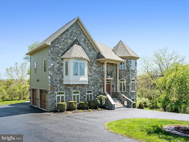 french country inspired facade featuring a garage, a balcony, aphalt driveway, and stone siding