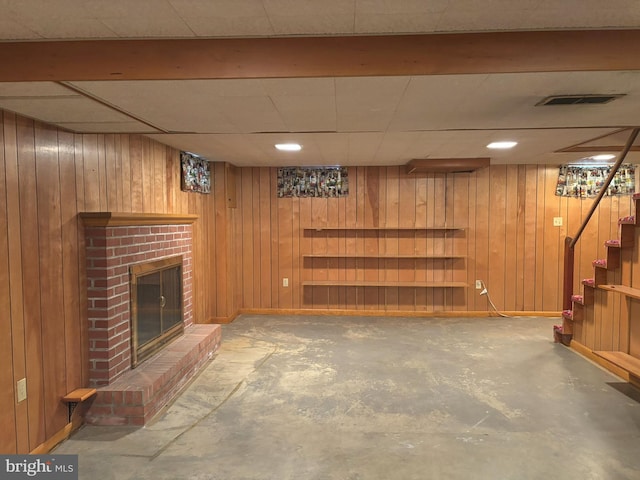 finished basement featuring wooden walls, baseboards, visible vents, stairs, and a brick fireplace