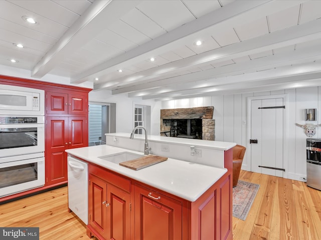 kitchen featuring white appliances, a center island with sink, a sink, a stone fireplace, and beamed ceiling