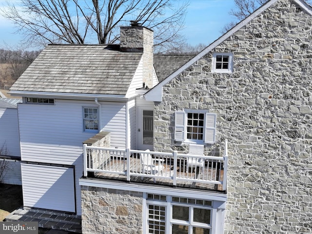 back of house with stone siding, a balcony, and a chimney