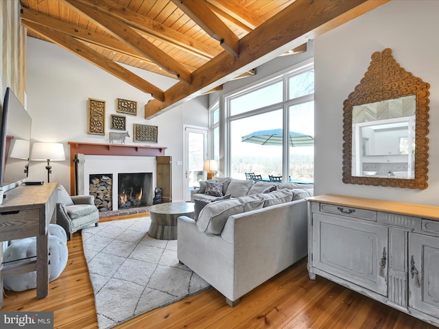 living area with beamed ceiling, high vaulted ceiling, a warm lit fireplace, light wood-style floors, and wooden ceiling