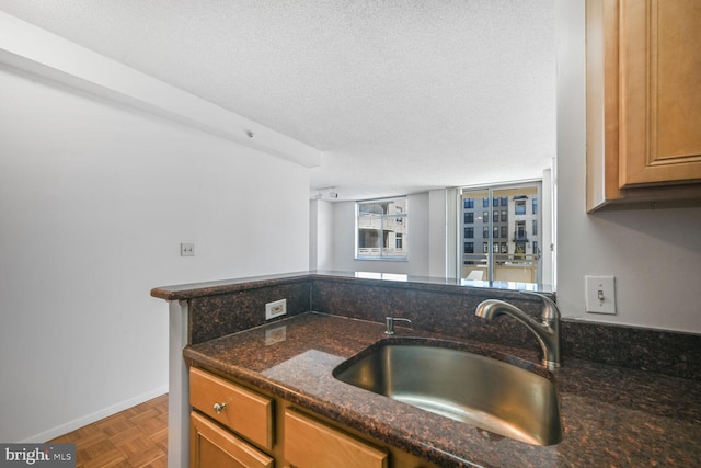 kitchen with brown cabinetry, a sink, a textured ceiling, dark stone countertops, and baseboards